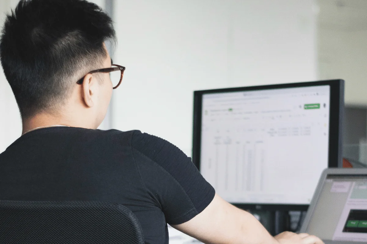 A person in glasses and a black shirt is working intently on a computer, with spreadsheets visible on the screen, in a bright, modern office setting.