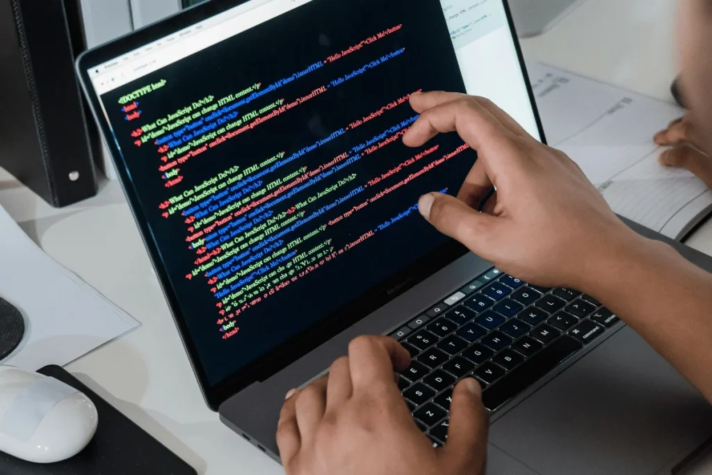Hands typing on a laptop displaying colorful HTML code. Intense focus on coding, with notes and a computer mouse visible nearby on a white desk.