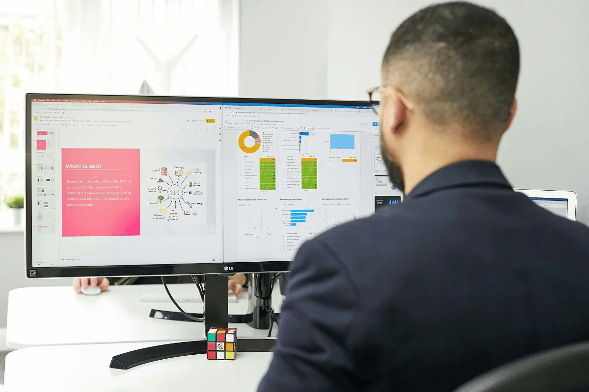 A man in a suit works at a dual-monitor setup, displaying charts and a presentation on SEO.
