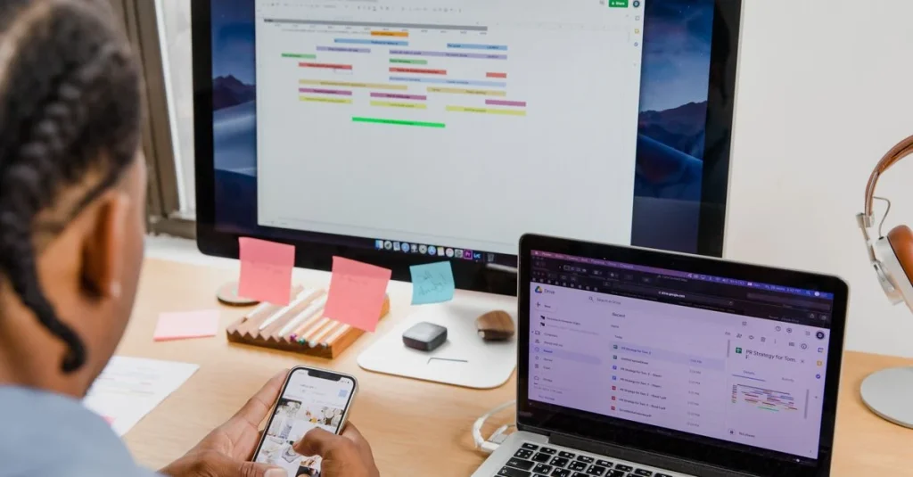 Person using smartphone at desk with business intelligence reporting dashboard displayed on monitor and laptop.