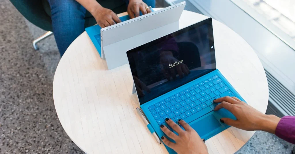 Person unboxing Surface device with blue keyboard on white table by window.